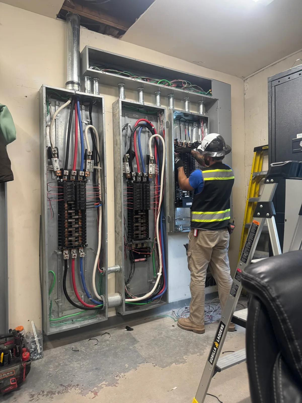 Electrician in hi-vis vest and hard hat wiring a bank of three commercial panel boards